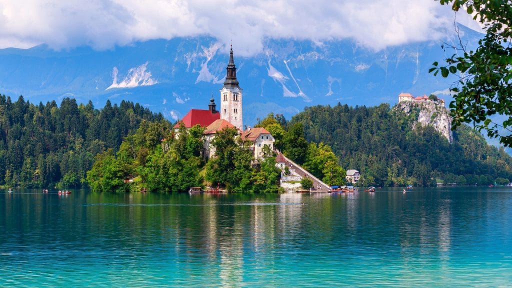 Bled Island with its church, bell tower and stone staircase above turquoise lake water, with a castle on a distant hill.