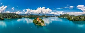 Aerial view of Bled Island with its church and bell tower, surrounded by the turquoise water of Lake Bled, wooded shores, hills and clouded mountains.
