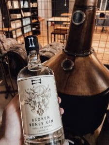 A hand holds a Broken Bones Gin bottle in front of a copper still inside the distillery.