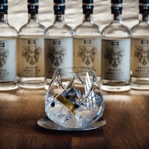 A gin glass with ice, lemon peel and juniper berries in front of Broken Bones Gin bottles.