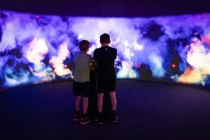 Two children seen from behind facing a large colorful projected space scene in a dark immersive room at Center Noordung.