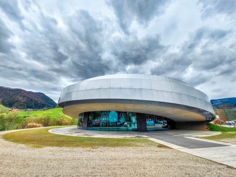 Circular silver-and-concrete Center Noordung building in Vitanje beneath a cloudy sky, with green hills behind.