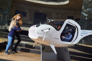 White pod-shaped flight simulator at Center Noordung with a child wearing a VR headset in the front seat and an adult seated behind, while another child and an adult stand beside the pod.