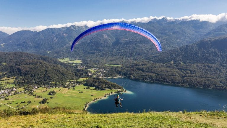 Tandem paraglider flying above Lake Bohinj from the Vogar side, with Alpine peaks behind.
