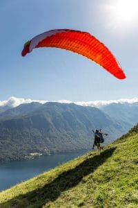 Tandem pilot and passenger running beneath a red paraglider on the Vogar launch slope above Lake Bohinj.