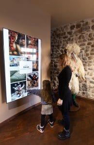 An adult and a child examine a wall display of colorful images beside a Kurent costume.