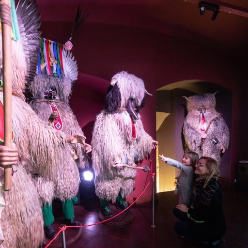 An adult crouches next to a small child who points at several full Kurent costumes in a pink-lit gallery.