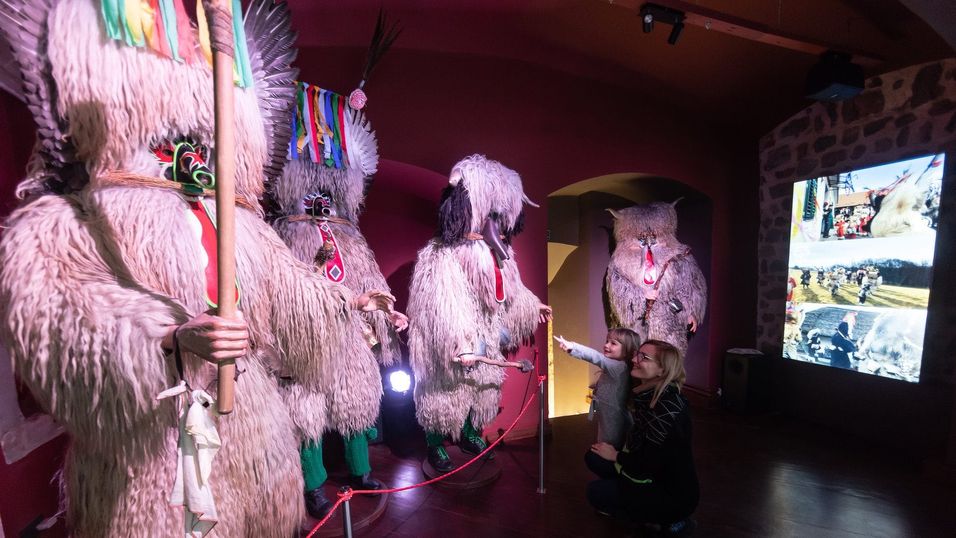 An adult crouches next to a small child who points at several full Kurent costumes in a pink-lit gallery.