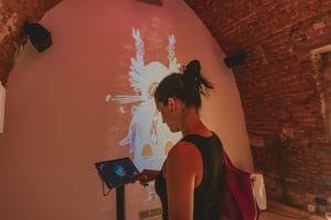 A visitor uses a tablet station beneath a wall projection of a Kurent figure.