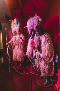 Two full sheepskin Kurent costumes with feathered headpieces stand behind a rope barrier.