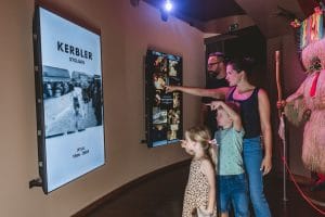 Two adults and two children study photographs beside an illuminated Kerbler display and a Kurent costume.