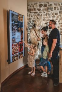 Two adults and two children look at photographs on a display beside a Kurent costume inside the house.