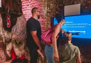 Two adults and two children gather at a bright blue interactive screen beside a sheepskin Kurent costume.