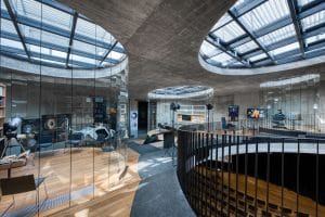Curving concrete interior at Center Noordung with circular skylights, glass walls and space exhibits around a central opening that looks down onto rows of seats below.