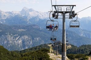 Riders on the open chairs of the Orlove glave chairlift, with the Julian Alps behind.