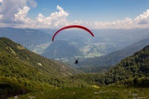 Paraglider flying under a red canopy between forested mountain slopes above the Bohinj valley.