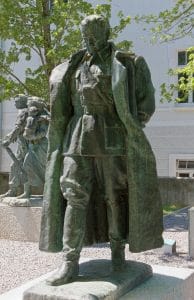 Two bronze sculptures on stone plinths outside a pale building: a uniformed figure in a long coat, and behind it a second group including a figure carrying a rifle.