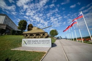 Tank above the bilingual Park of Military History entrance sign, with Slovenian flags lining the road.