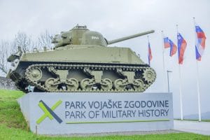 Tank mounted above the bilingual entrance sign at Park of Military History Pivka, with Slovenian flags behind it.