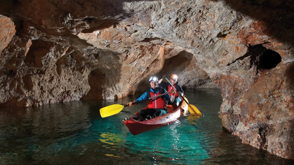 Two helmeted paddlers in a red kayak move through flooded mine tunnels.