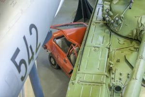 A crushed red car displayed between a military aircraft fuselage and a green armoured vehicle inside the Pivka military museum.