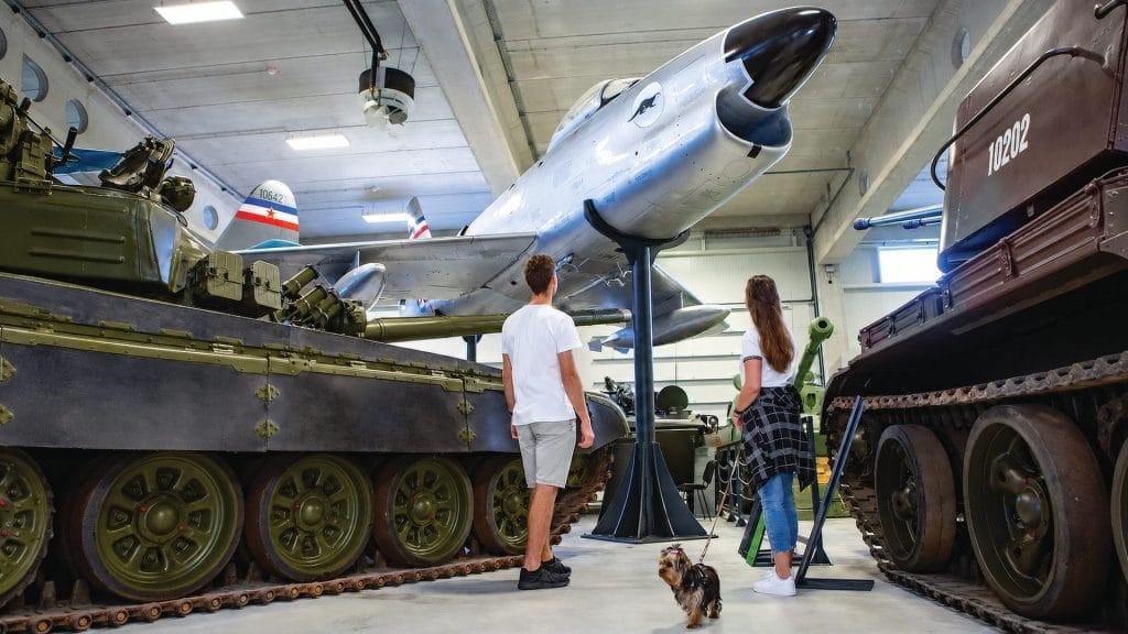Two visitors with a small dog on a leash standing between armoured vehicles beneath a historic aircraft in the museum hall.
