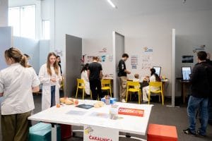 People, several in white coats, sit and stand at clinic-themed stations inside Tehnopark Celje.