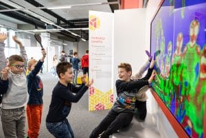 Children raise their arms and touch an infrared camera display showing colourful silhouettes at Tehnopark Celje.