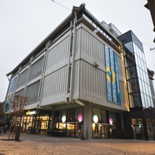 The Tehnopark Celje building with vertical banners and a glass corner stands on a pedestrian street at dusk.