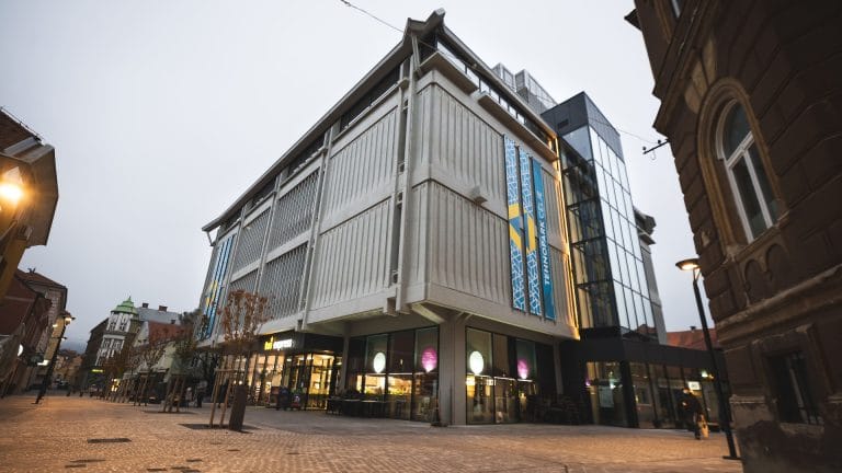 The Tehnopark Celje building with vertical banners and a glass corner stands on a pedestrian street at dusk.