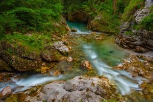 A clear stream runs over rocks in front of a turquoise pool between wooded stone banks.