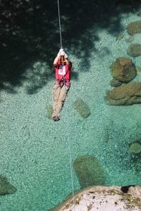 Zipline rider seen from above over clear turquoise water and rocks in Učja Canyon.