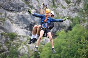 Two helmeted riders descending side by side on parallel zipline cables above Učja Canyon.