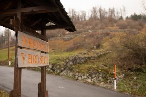 Wooden Welcome to Hrušica sign beside the road with Roman wall remains on the slope.