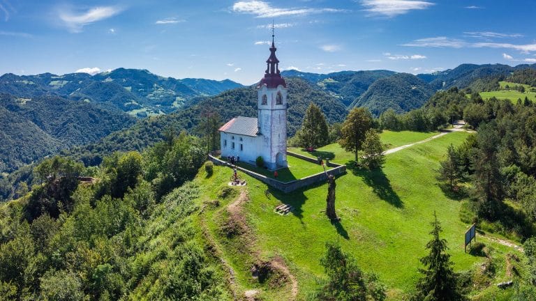 Aerial view of a white church with a red-roofed bell tower and a walled churchyard on a grassy ridge above wooded hills.