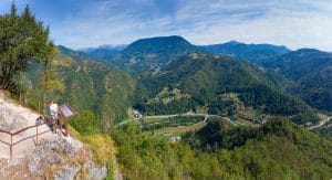 A small group with a young child and a dog at a railing overlooking a winding valley and wooded mountains near Divje Babe.