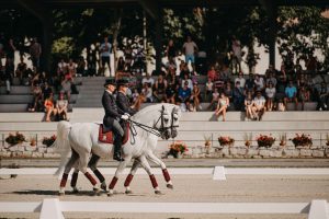 Two riders in dark tailcoats and top hats ride a pair of white horses side by side across an outdoor arena, with spectators behind.