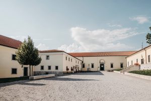 Pale historic buildings surround a broad gravel courtyard with a stone well under a bright sky.